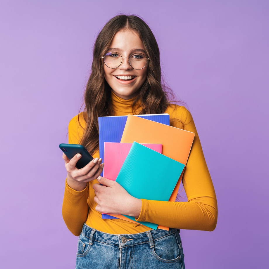 Eine junge Frau mit Brille hält Bücher und ein Handy und lächelt.
